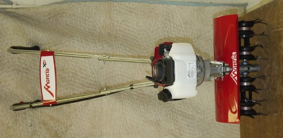 A red and white Mantis tiller with handles lying on a beige surface.