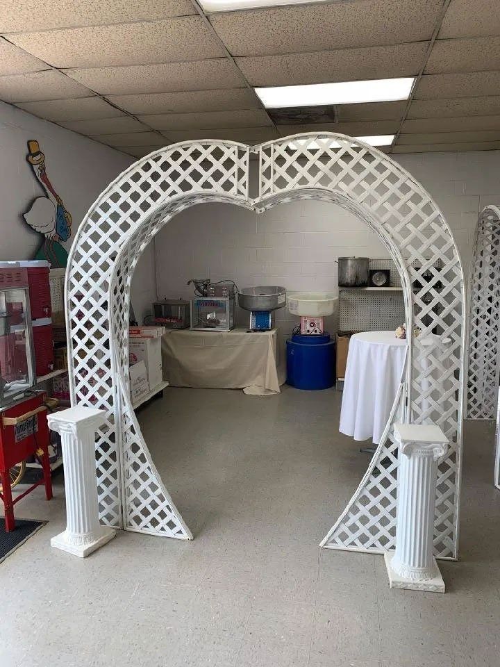 White heart-shaped trellis archway with pillars indoors; party equipment visible in the background.