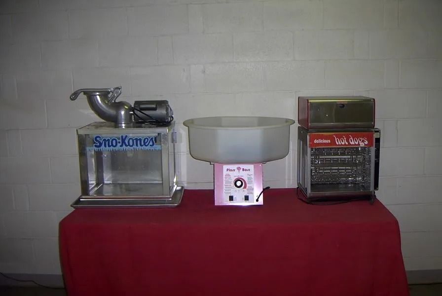 Three carnival food machines on a red table: snow cone, cotton candy, and hot dog roller.