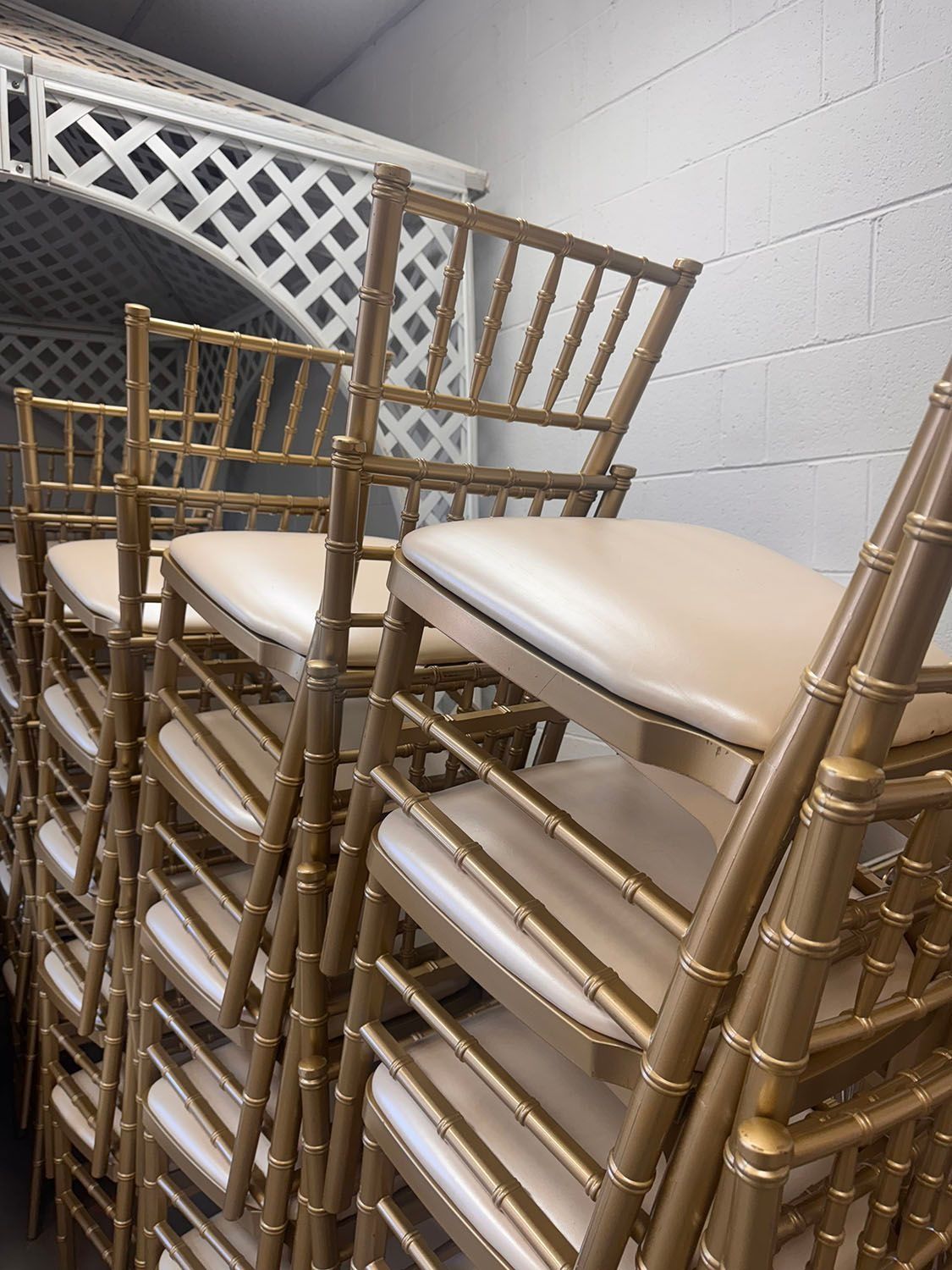 Gold Chiavari chairs stacked near a white lattice structure. Beige cushions are visible.