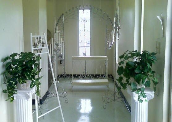 Hallway with potted plants, ornate metal arches, and a window with a simple altar in the background.
