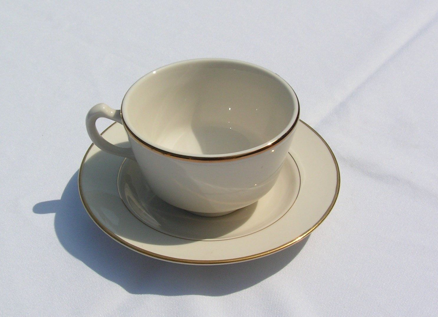 Cream-colored teacup and saucer with gold trim, sitting on a white tablecloth.