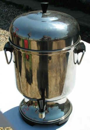 Shiny, stainless steel coffee urn with lid, handles, and spigot, outdoors.