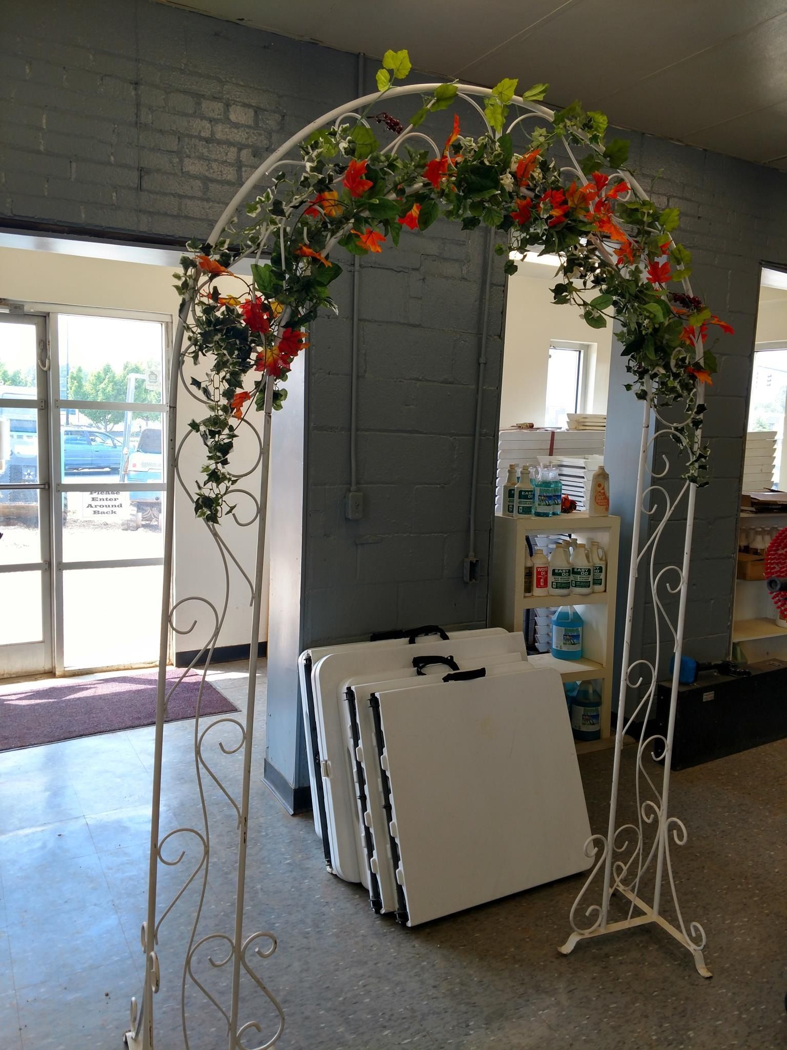 White metal arch decorated with orange flowers, near folded white tables, and glass doors.