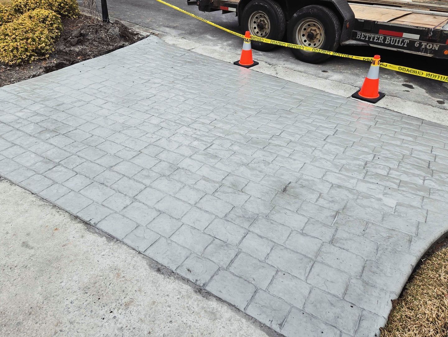 Newly poured concrete patio with brick pattern; caution tape and orange cones.