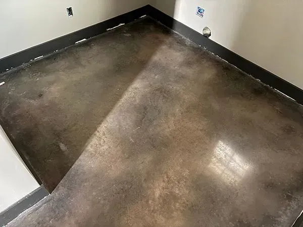 Brown stained concrete floor in a corner of a room, with dark trim and unfinished walls.