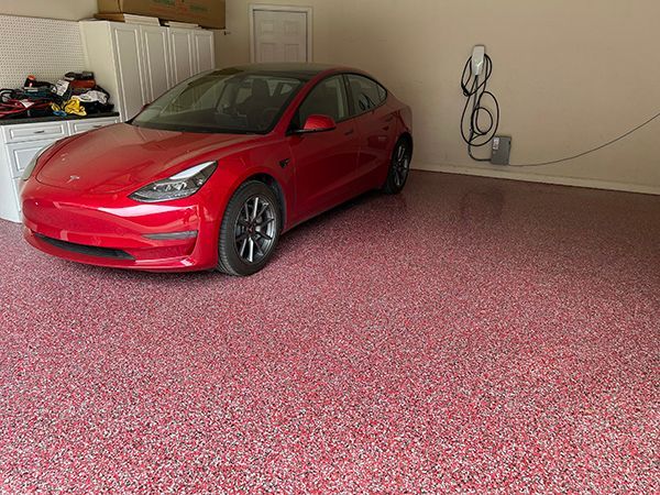 Red Tesla in a garage with a red flecked floor, charging station on the wall, and white cabinets.