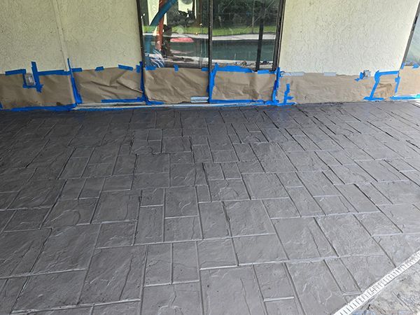 Stamped concrete patio with a gray finish, taped off near a wall and sliding glass door.