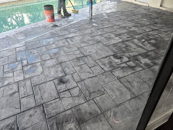 Gray stamped concrete patio next to a pool. A person is using a broom on the wet surface.