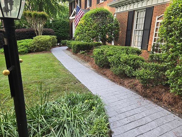 A gray brick pathway leads to a brick house with black shutters and an American flag. Green bushes line the path.