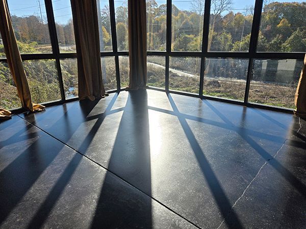 Sunlight streams through tall windows onto a dark, glossy floor, illuminating a corner room with curtains and a view.