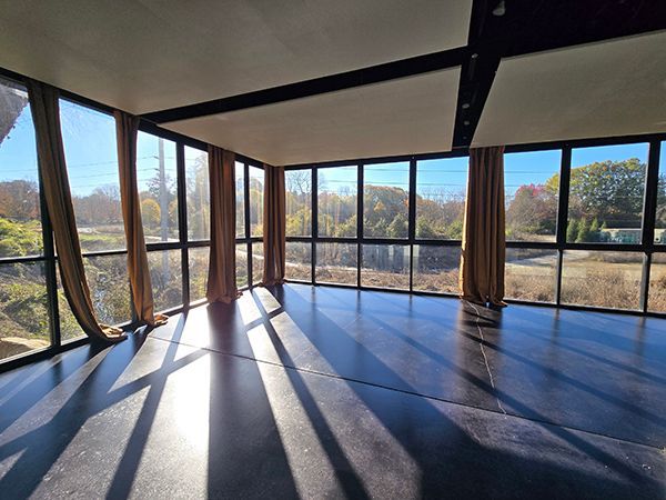 An empty room with floor-to-ceiling windows, dark floor, and curtains. Sunlight creates shadows.