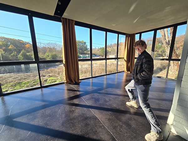 Person in a room with large windows looking outside, wearing a jacket and jeans.