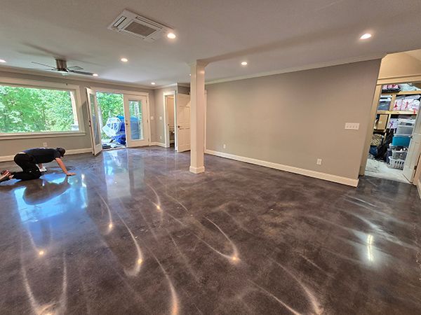 A person cleaning a shiny, gray epoxy floor in a spacious room with large windows and a neutral-colored wall.
