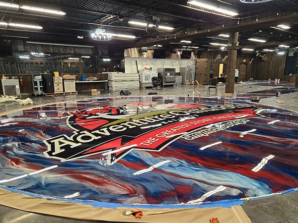 Large floor mural in progress; Adventure Cove logo in red, white, and blue on a swirled epoxy floor.