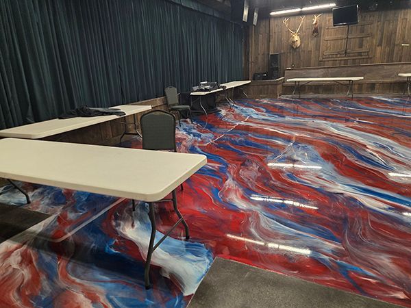 A room with red, white, and blue swirled epoxy floor, white tables, dark curtains, and wood paneling.