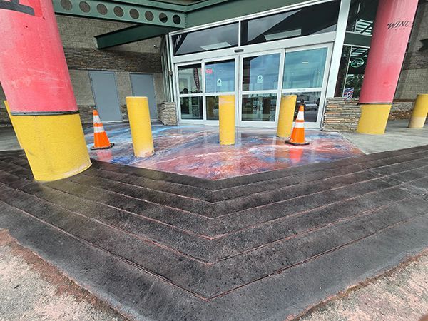 Exterior entrance with steps, red/yellow columns, decorative pavement. Orange traffic cones are placed around.