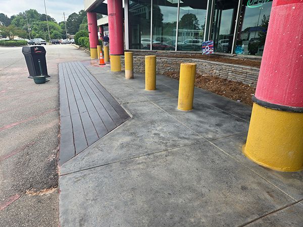 Sidewalk with ramp, yellow bollards, red and yellow pillars. Black trash can.