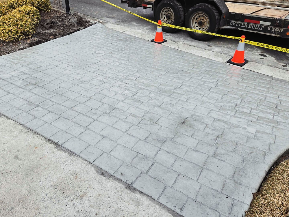 Stamped concrete pathway, gray, with a brick pattern. Orange cones and a trailer are in the background.