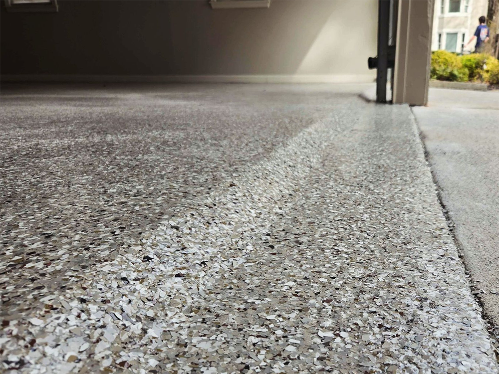 Epoxy garage floor with gray, white, and tan chips, next to a concrete driveway and a partially open garage.