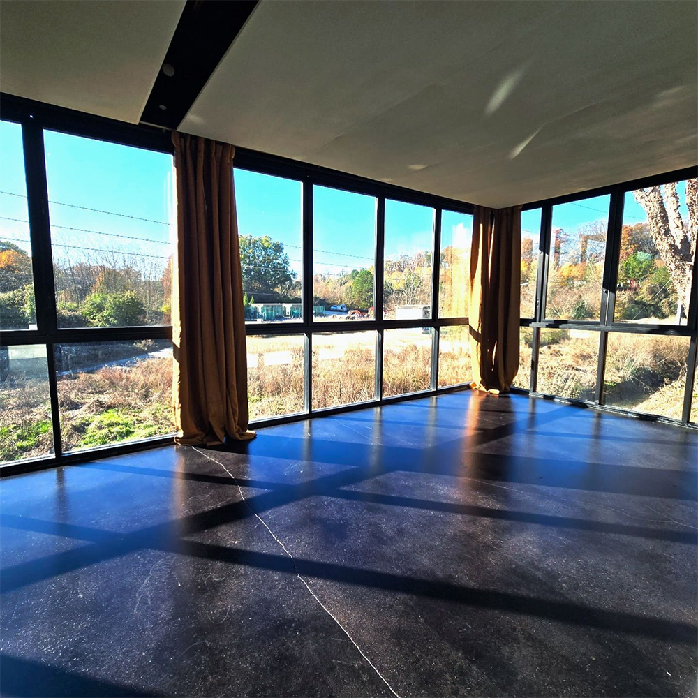 Interior view of a room with large windows overlooking an autumn landscape. Dark floor, tan curtains.