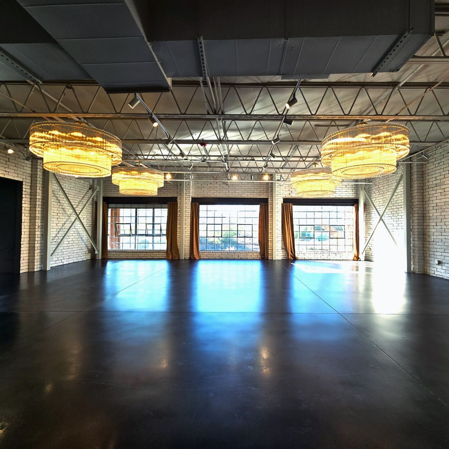 Empty modern event space with dark floor, brick walls, large windows, and unique hanging lights.