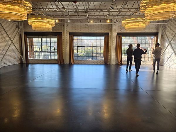 Empty industrial event space with three figures near windows, gold chandeliers above.
