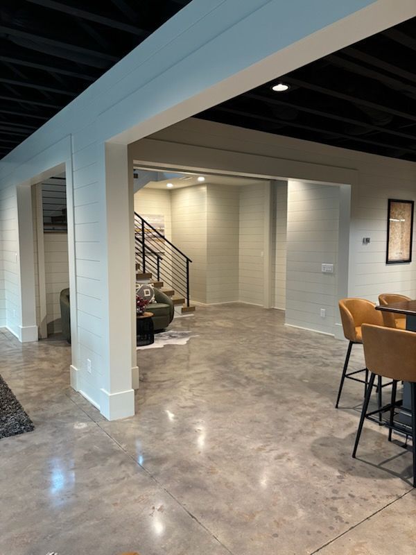 Spacious basement with concrete floor, white walls, black ceiling, staircase, and bar stools.