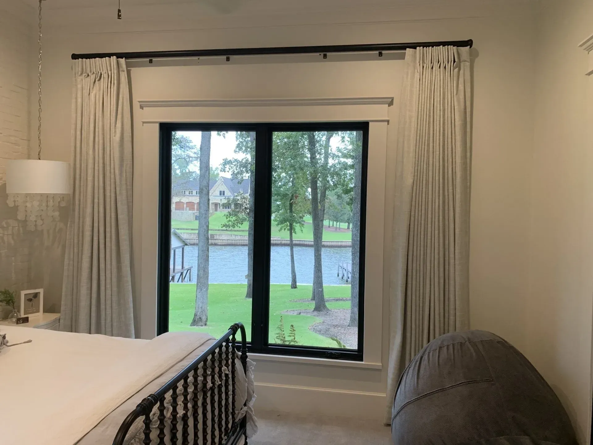 Bedroom with bed, black metal headboard, floor lamp, and a large window overlooking trees and water.