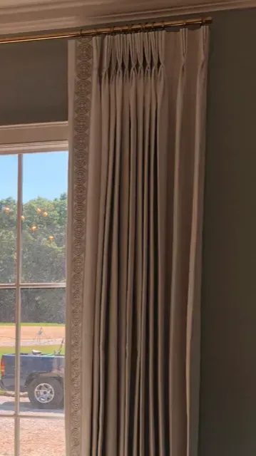 Beige pleated curtains beside a window with a view of parked cars and trees outside.