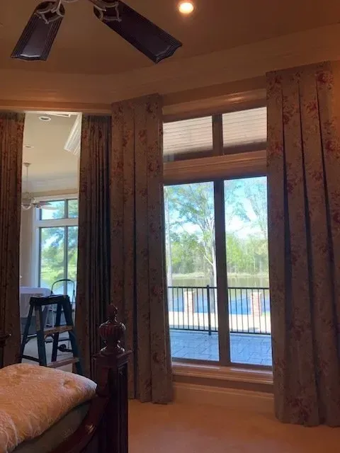 Bedroom with a large window, curtains, and a ceiling fan, overlooking a balcony and greenery.