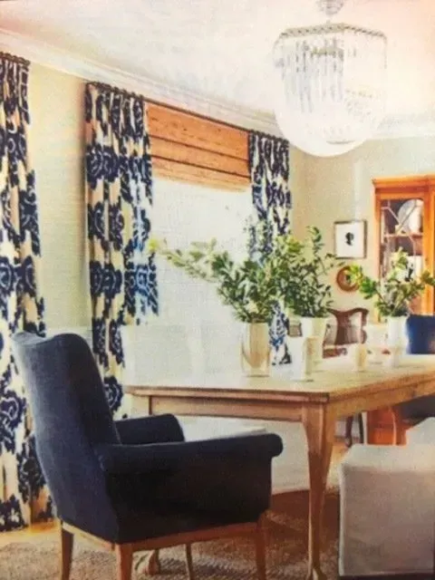 Bright living room with a wooden table, dark armchair, white chair, curtains, and potted plants by a window