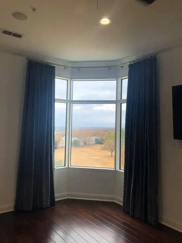Empty room with dark wood floors, blue curtains, and a bay window overlooking a field