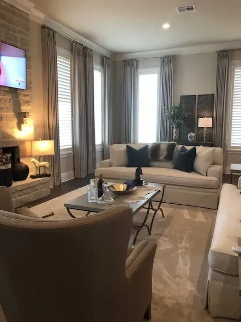 Cozy living room with beige sofa, armchair, glass coffee table, and tall windows with curtains.