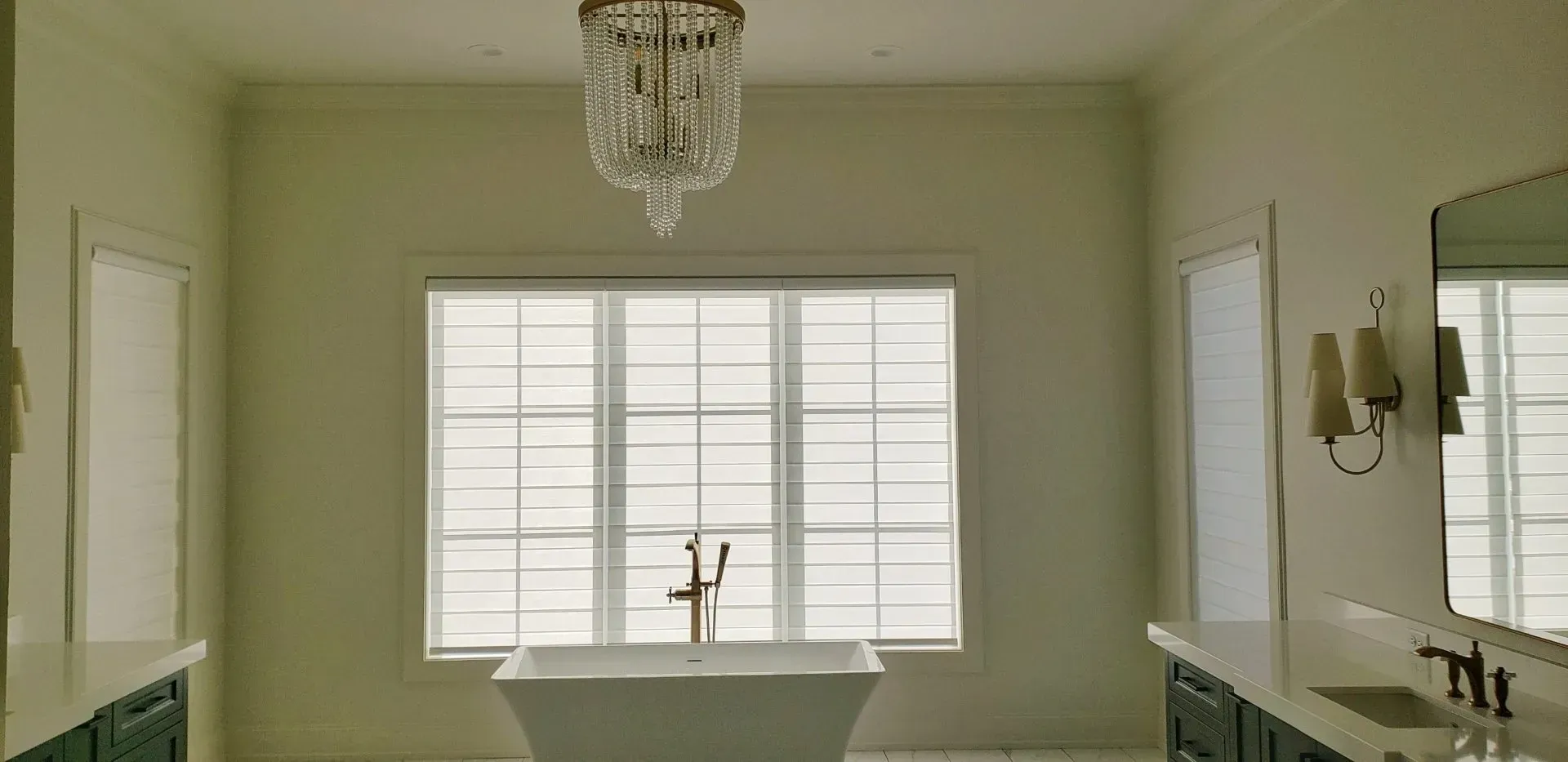 Bright bathroom with a centered tub under a window, chandelier, and large wall mirror.