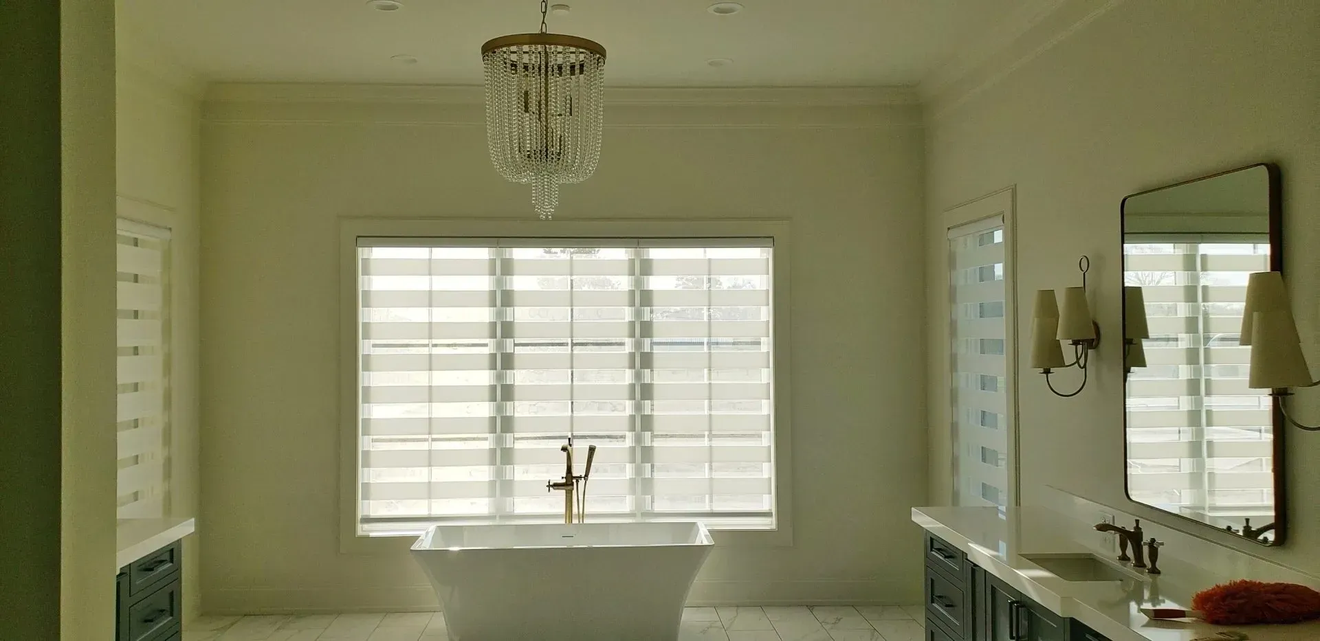 Bright bathroom with window blinds, bathtub, chandelier, and mirror above a vanity