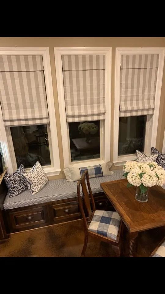 Cozy window nook with cushioned bench, wooden table, plaid chair, and flowers by three curtained windows