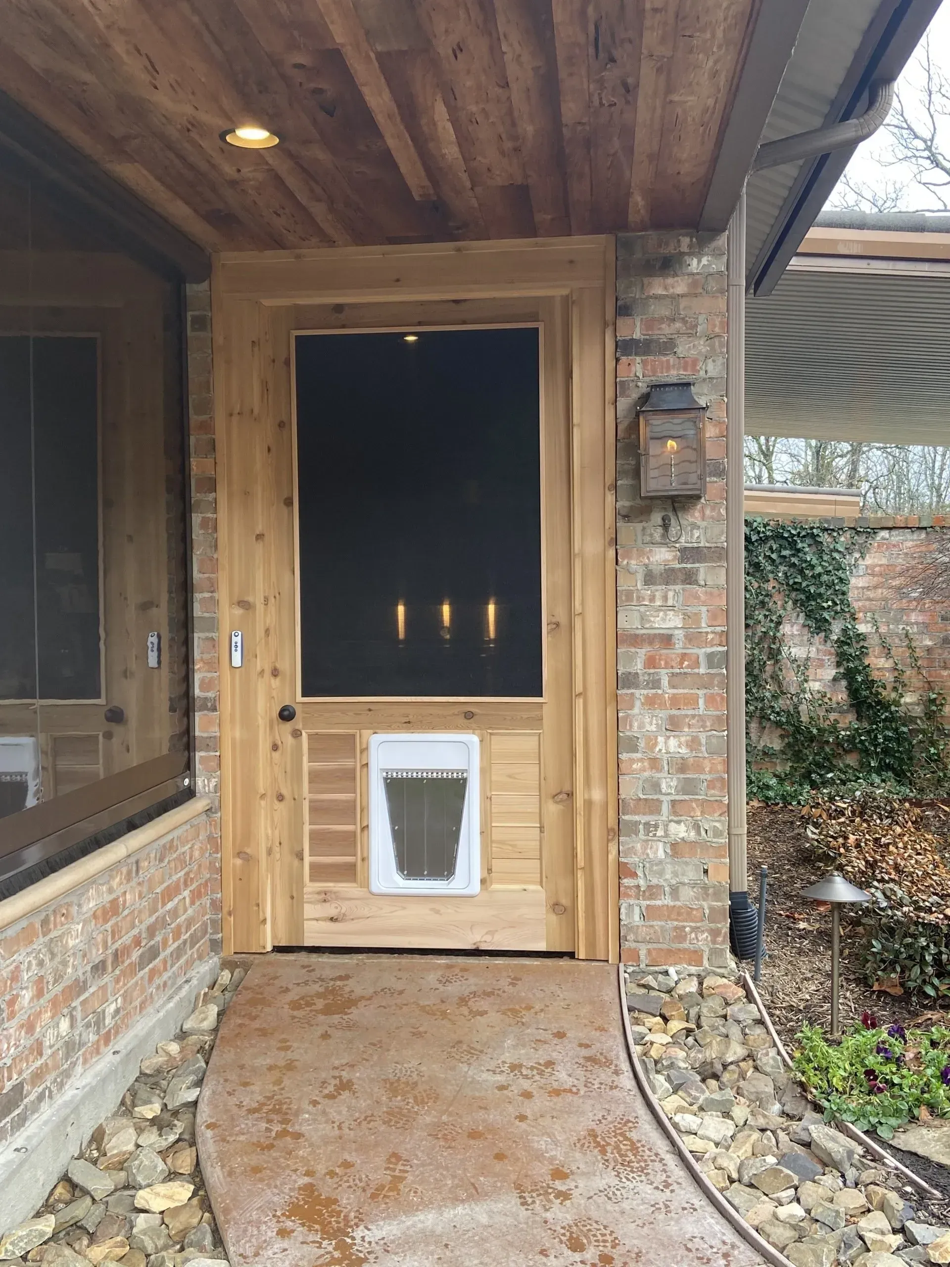Wooden front door with a pet flap under a covered brick porch, beside a window and brick wall.