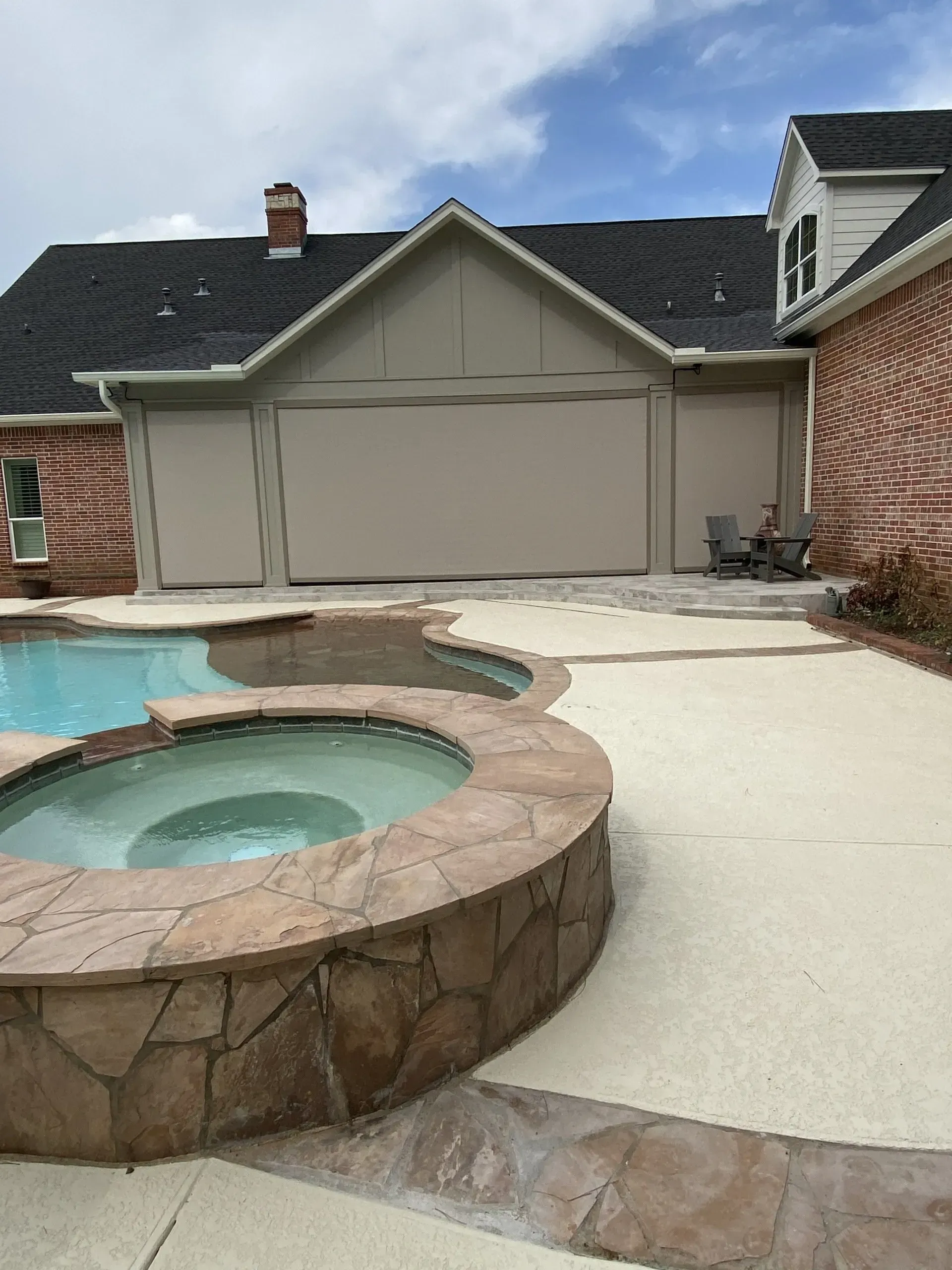 Backyard patio with stone hot tub, swimming pool, and beige garage beside a brick house