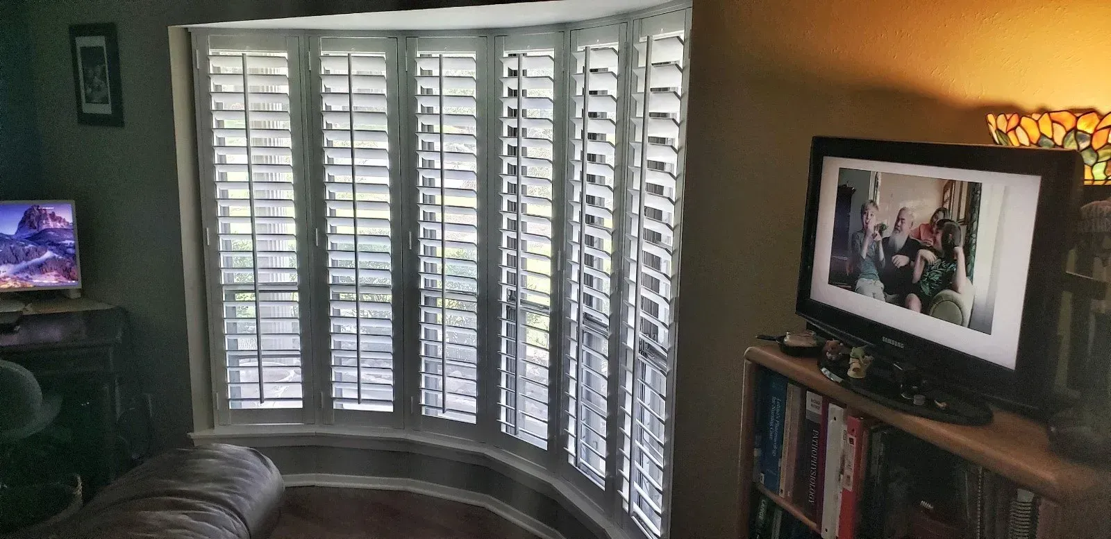 Living room with white shutters, a TV on the left, and a framed photo on a shelf by the window.