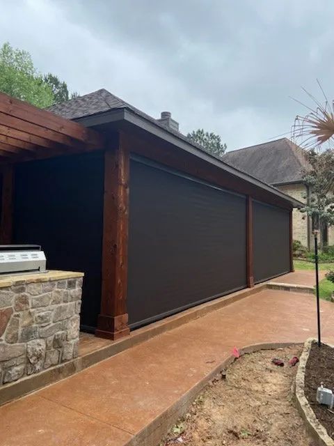 Covered patio with dark roll-down shade and stone columns beside a wet walkway and landscaped yard