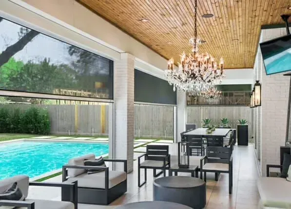 Covered patio dining area beside a swimming pool, with black furniture, chandelier, and wood ceiling.