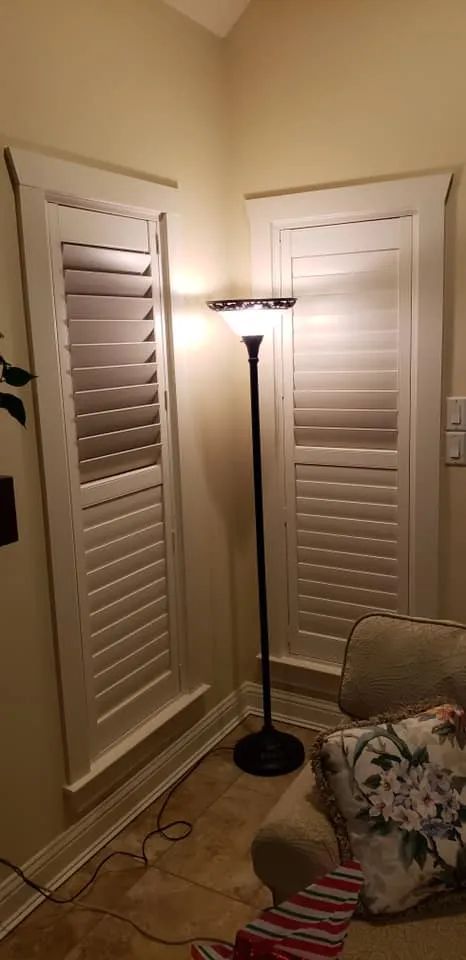 Corner of a room with white plantation shutters, a black floor lamp, beige tile, and a floral cushion