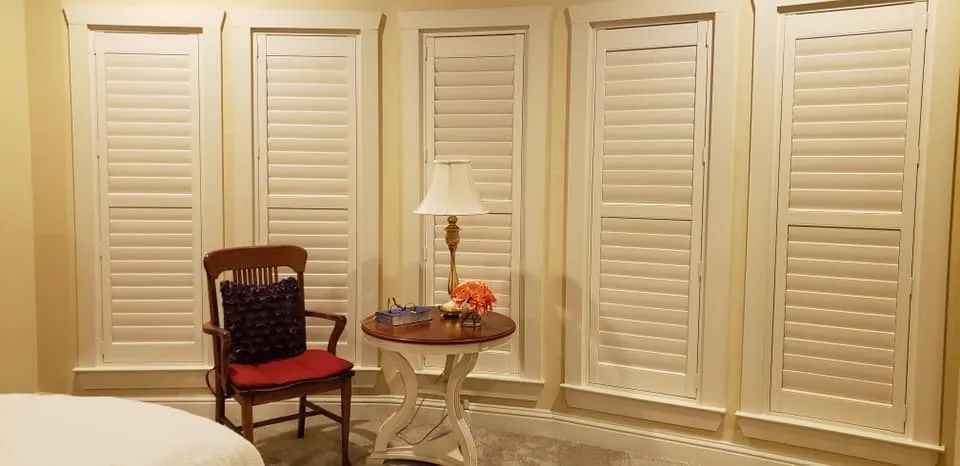 Cream-colored room with white shutters, a red chair, and a small table with lamp and fruit bowl.