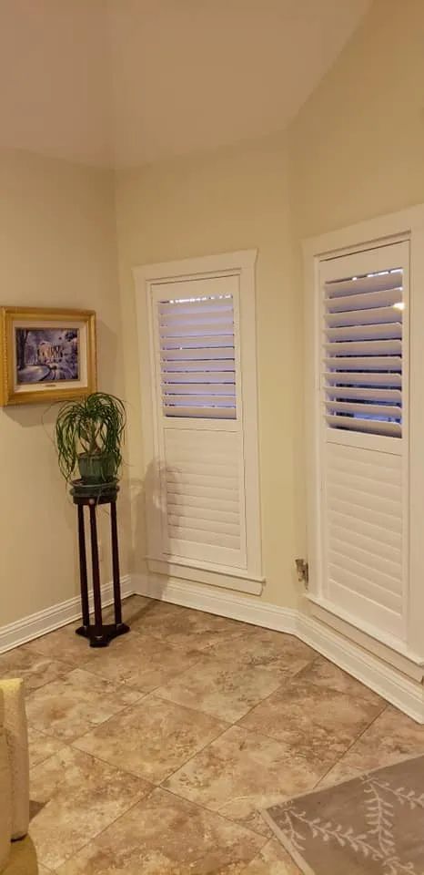 Empty beige corner with two white shuttered windows, a plant on a stand, and a framed picture.