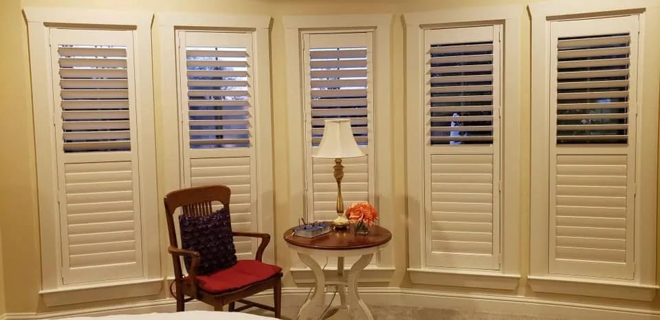 Sunlit bay window with white shutters, a red chair, small table, lamp, and orange flowers