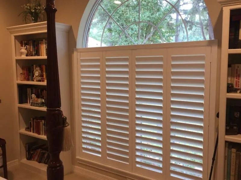 White plantation shutters cover a large arched window in a room with built-in bookshelves.