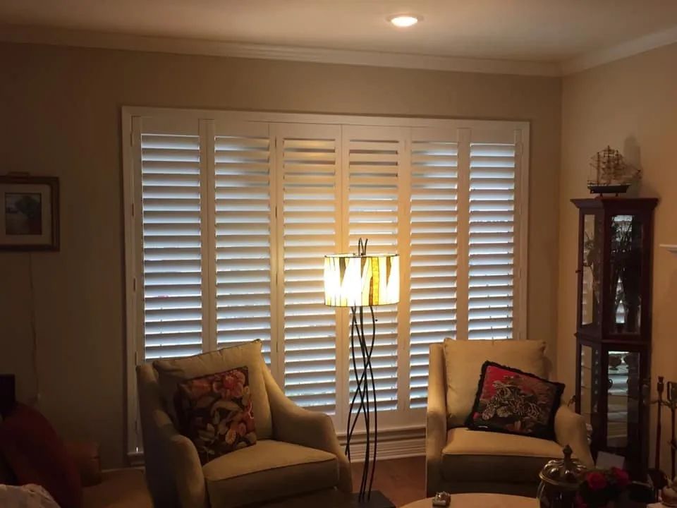 Living room with window shutters, two chairs, and a floor lamp beside a glass display cabinet