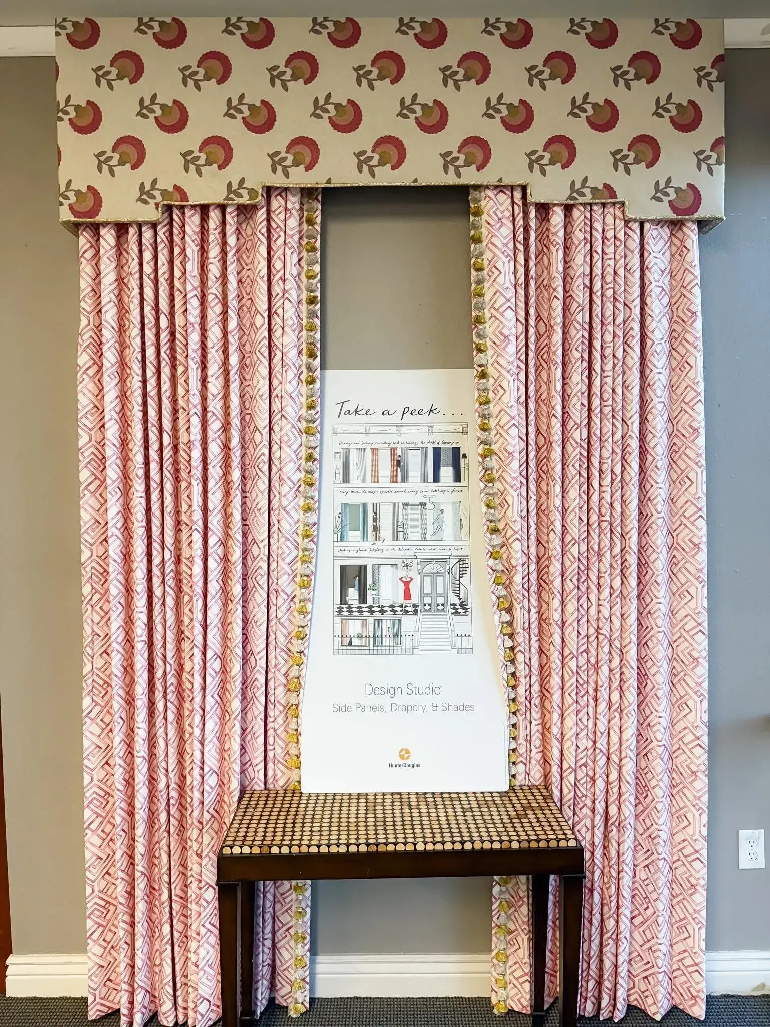 Pink patterned curtains frame a display board behind a small wicker table in a room.