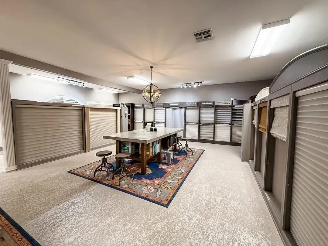 Spacious indoor room with beige walls, multiple window blinds, central table, and patterned rug.